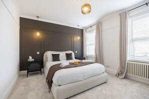 A modern bedroom with a large bed, bedside tables, a hanging light fixture, and a window with curtains. - Stylish 2-Bed Home Close to Cheltenham Town Centre (Cheltenham)