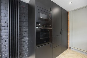 A kitchen with a microwave, oven, and built-in refrigerator, adjacent to a brick wall and a white door. - Stylish 2-Bed Home Close to Cheltenham Town Centre (Cheltenham)