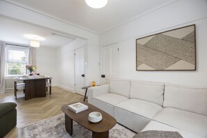 En moderne stue med en hvid sofa, et træsofabord, et spisebord med stole og et stort abstrakt maleri på væggen.