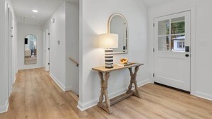 Un pasillo con suelo de madera, una puerta blanca con ventanal y una mesa con lámpara y adornos.