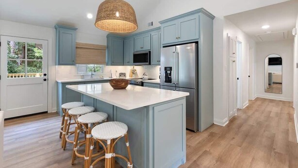 Una cocina con alacenas azul claro, una isla grande con taburetes, una lámpara colgante y un refrigerador.