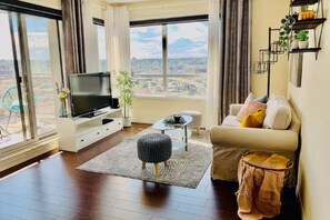 A modern living room with a large window, a flat-screen TV, a sofa, a coffee table, and a small side table. - New | Intro Price | Cozy 950sf 2BR+2BA | AC &Gym &Parking | City &Mountain View (Calgary)