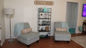 Uma sala de estar com dois poltronas estampadas, um abajur, uma estante com objetos decorativos e uma placa com a inscrição 'BEACH HOUSE'.