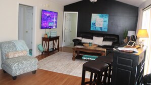 Uma sala de estar com parede preta, ventilador de teto, televisão, sofá, mesa de centro e escrivaninha com luminária.