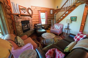 A log cabin interior with a stone fireplace, leather sofas, a staircase, and a view of the outdoors. - Peaceful Lakeside Cabin on Minerva Lake-Camp 16 (Olmstedville)