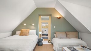Un dormitorio con dos camas, una mesita de noche con lámpara, una pequeña mesa con un libro y vistas a un baño a través de una puerta.