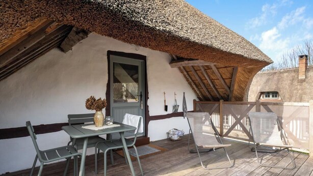 Una casa rústica con techo de paja y una terraza de madera, una mesa con sillas y un jarrón de flores.
