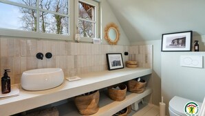 Un baño moderno con un lavamanos, tocador y retrete blancos. Hay cestos de mimbre debajo del tocador, un cuadro en la pared y una ventana con vistas a árboles.