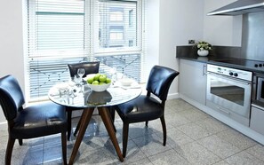 Eine moderne Küche mit Essbereich, darunter ein runder Glastisch mit Stühlen, eine Schüssel mit grünen Äpfeln und ein Edelstahlbackofen.