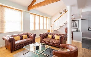 Ein geräumiges Wohnzimmer mit Ledersofas, einem Glastisch und einer Treppe, die zur Küche führt.