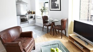 Een moderne keuken met roestvrijstalen apparatuur, een eethoek met twee stoelen, een glazen salontafel met appels en een televisie op een houten standaard.