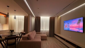 Un salon moderne comprenant un canapé d’angle, une table à manger avec des chaises, un téléviseur à écran plat fixé au mur et une grande fenêtre avec des rideaux.