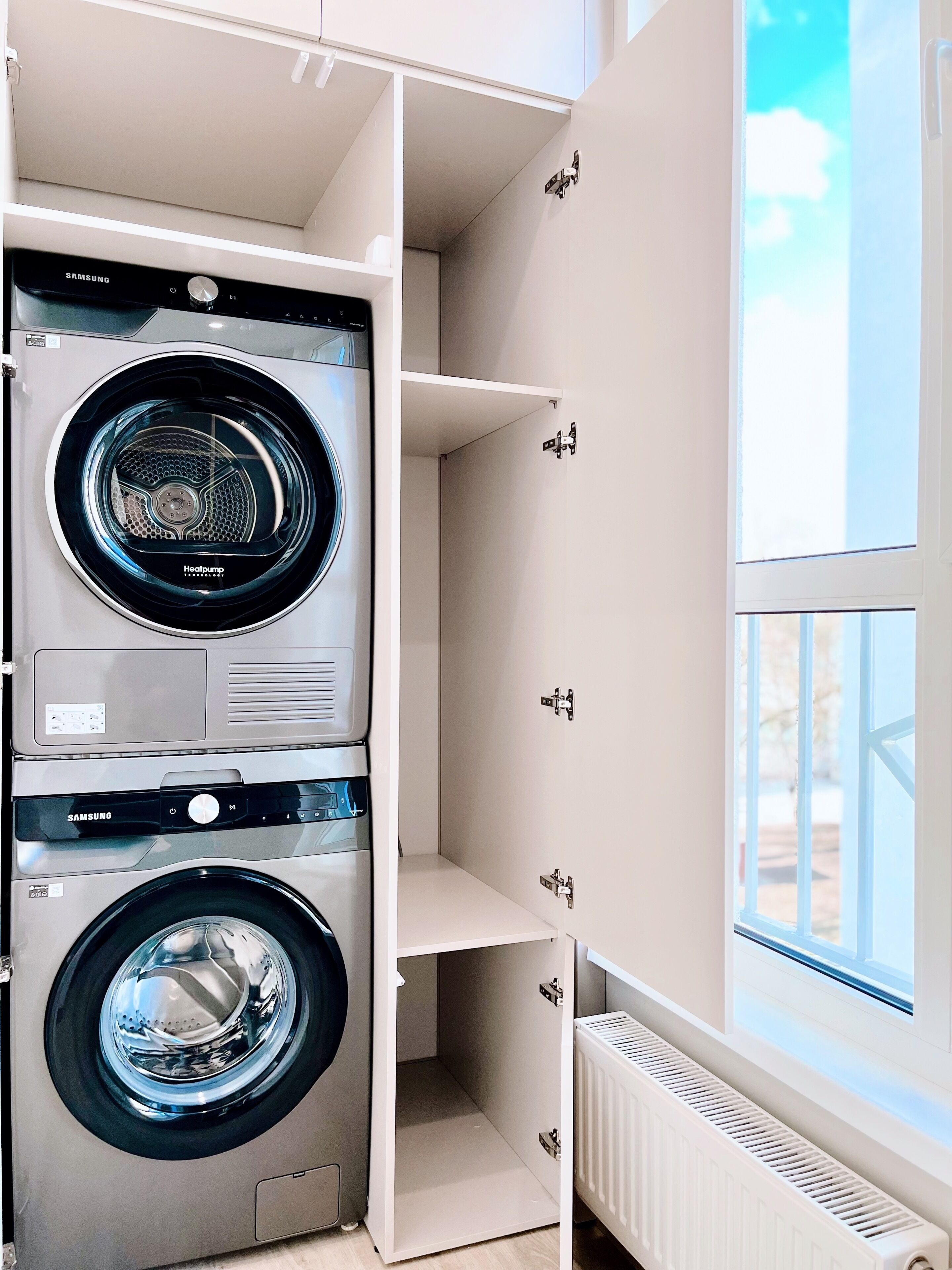 A laundry room with two Samsung washing machines, a radiator, and a window with a view.