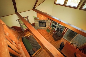 Ein Loft-Zimmer mit Kamin aus Stein, Holzbalken, einer grünen gepolsterten Bank und Blick ins Freie.