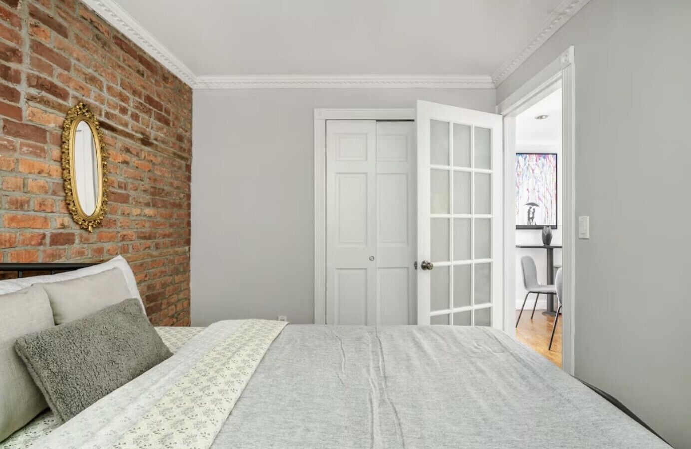 A bedroom with a brick wall, a white door, a bed with grey bedding, and a mirror.