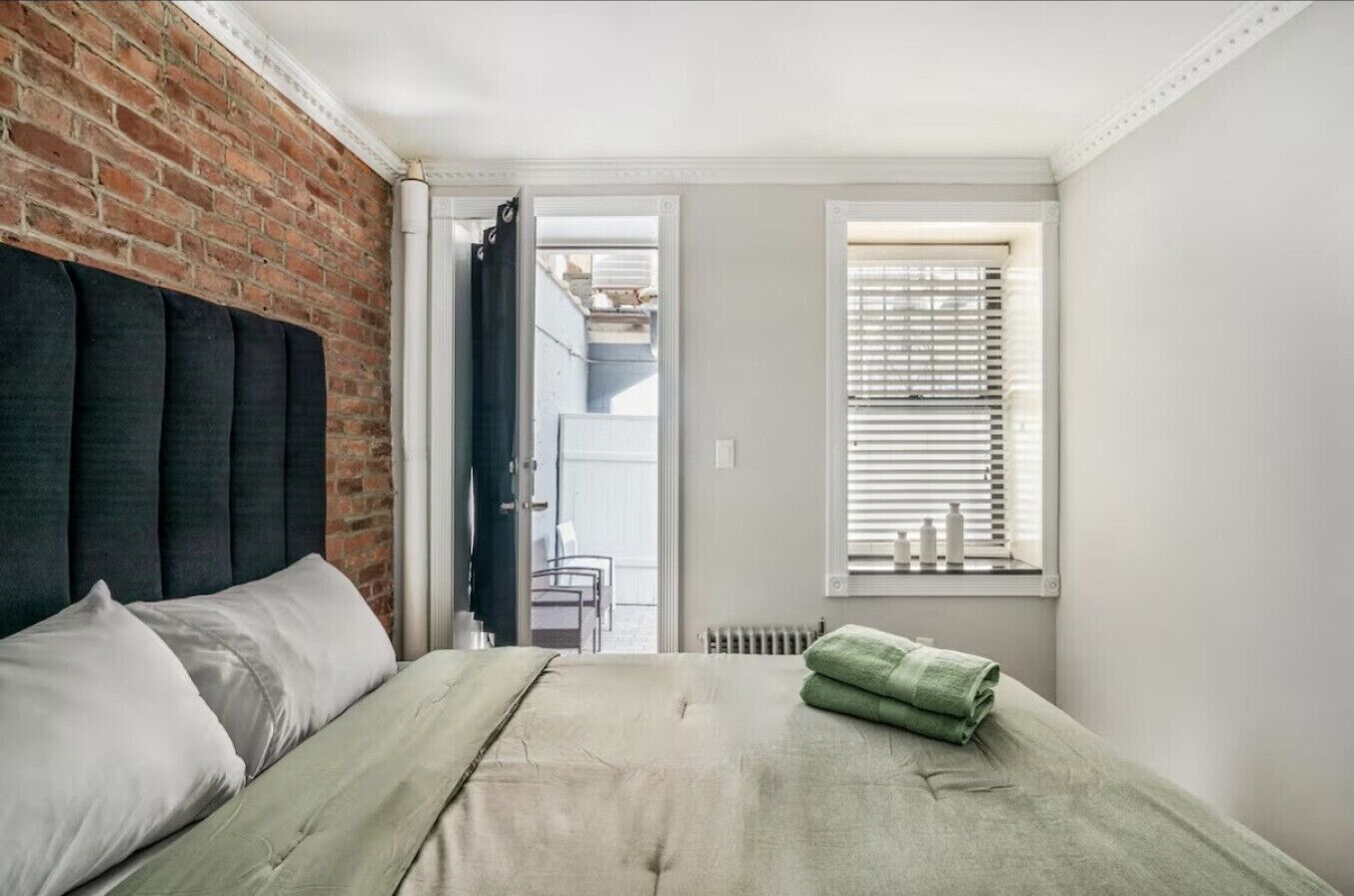 A bedroom with a brick wall, a bed with white and green bedding, a radiator, and a window with blinds.