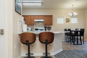 A kitchen with wooden cabinets, a white countertop, two bar stools, a dining table with chairs, and a chandelier.