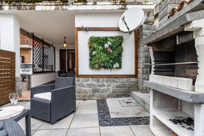 Un pati exterior amb mobiliari de vimet, una paret decorativa amb vegetació, una antena parabòlica i una llar de pedra.