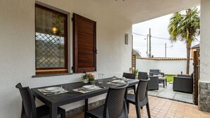 Een buitenplaats met een eettafel voor vier personen, omringd door rieten stoelen, een raam met houten luiken en uitzicht op een woonwijk.