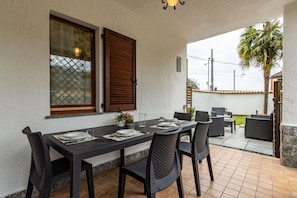 Un espace de restauration en plein air avec une table dressée pour quatre personnes, entourée de chaises en osier, une fenêtre avec des volets en bois et une vue sur un quartier résidentiel.