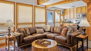 A cozy living room with a sectional sofa, a coffee table, and a kitchen area visible through an open door. - Fireplace, Deck, Walk to Slopes, Games, Views (Beech Mountain)