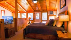 Ein Schlafzimmer mit Holzboden, einem Bett mit vielen Kissen, einem Fernseher links und einem Fenster mit Jalousien.