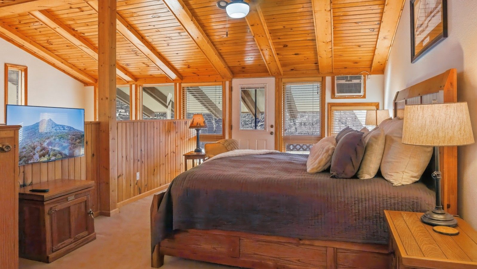 A bedroom with a wooden ceiling, a bed with pillows, a nightstand, a lamp, and a television on a wooden cabinet.