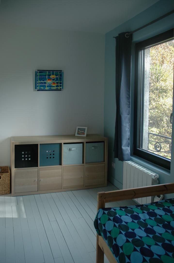 Ein Zimmer mit einem Holzkasten, einem Bett mit gemustertem Überwurf, einem Fenster mit Vorhängen und einem an der Wand hängenden Bild.
