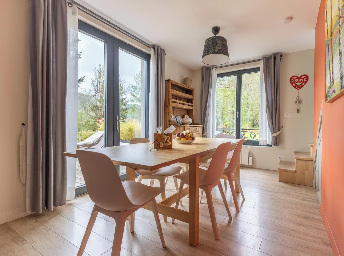 Un coin repas avec une table et des chaises en bois, de grandes fenêtres, une bibliothèque et un luminaire au plafond.