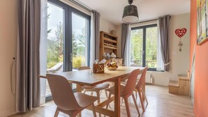 A dining area with a wooden table and chairs, large windows, a bookshelf, and a ceiling light fixture. - House La Couze - CHAMBON SUR LAC (CHAMBON SUR LAC)