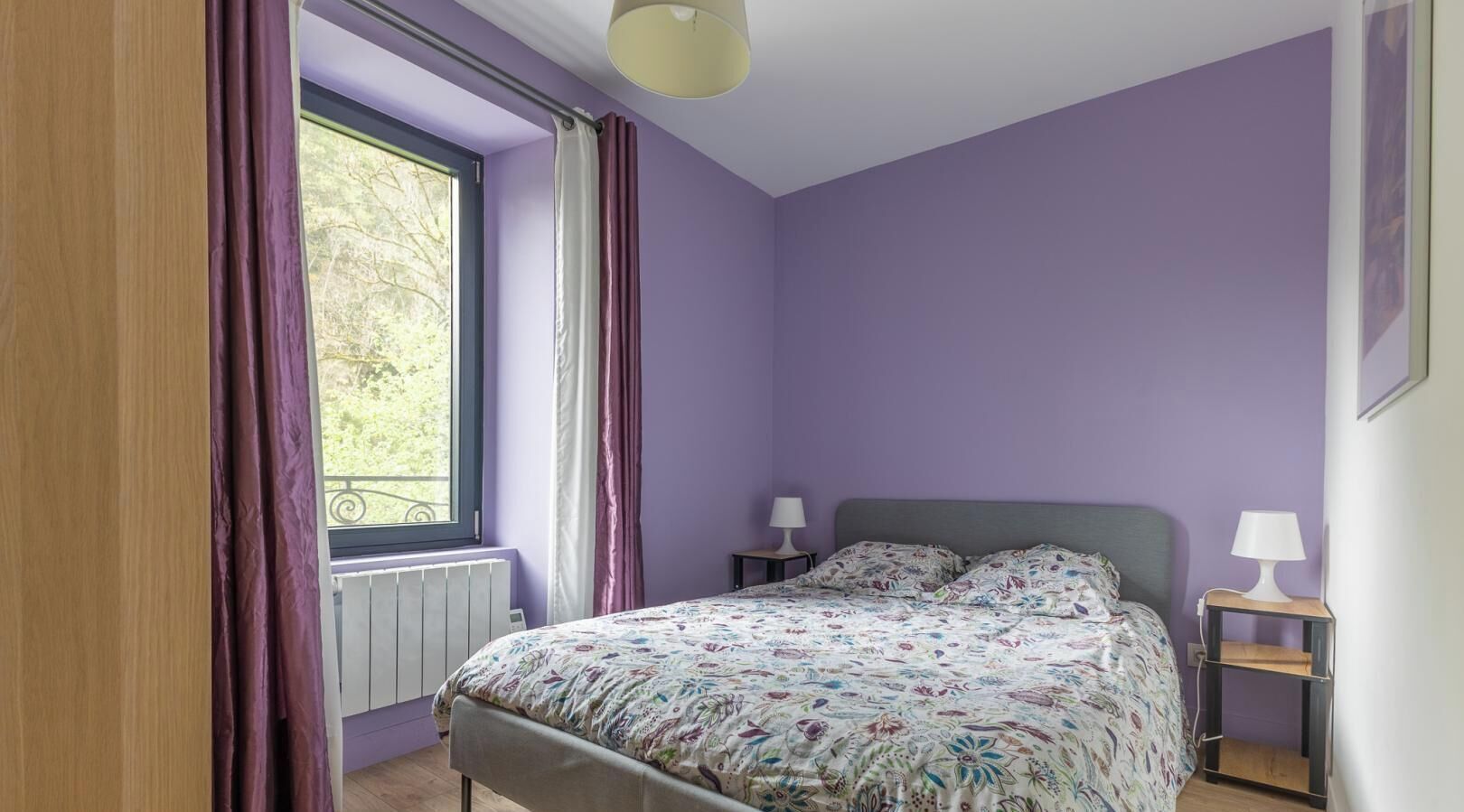 A bedroom with a bed, two bedside tables with lamps, a window with curtains, and a radiator.