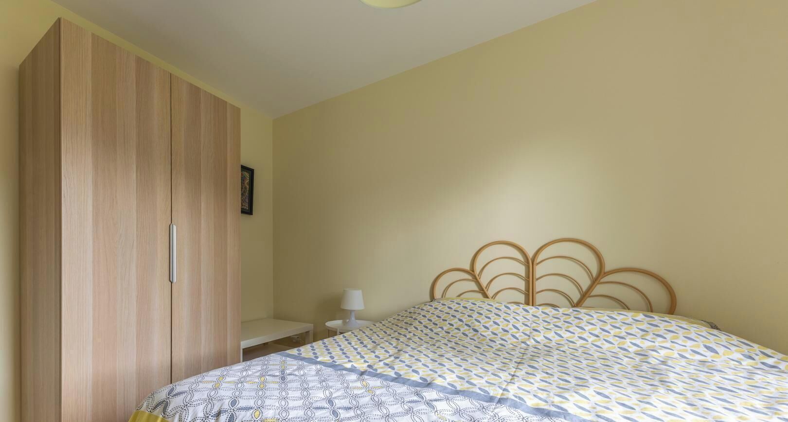 A bedroom with a bed, a wooden wardrobe, a bedside table with a lamp, and a wall-mounted picture.