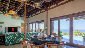 A dining area with a table set for six, surrounded by wicker chairs, a green patterned sofa, and a television displaying the Netflix logo. - Chalet with 3 bedrooms and panoramic views (Karangasem)