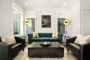 Un salon avec un canapé vert, deux fauteuils en cuir noir, une table basse noire et deux lampadaires.