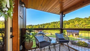 A wooden deck with two chairs and a table, overlooking a green landscape and a building. - Smoky Mountain Cabin: Hot Tub, BBQ & Fire Pit (Sevierville)