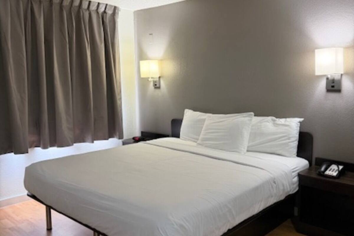 Ein Hotelzimmer mit einem Bett, Holzboden, einem Fenster mit Vorhängen und Wandlampen.
