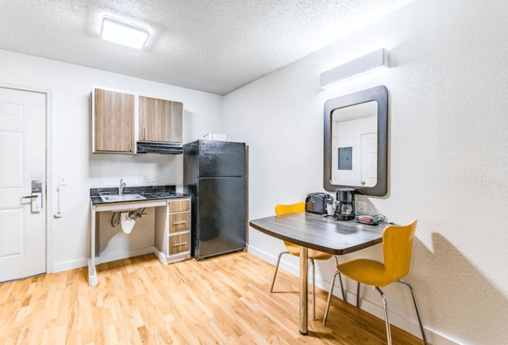 Cocina compacta con refrigerador, lavamanos y gabinetes de madera. Se ve una mesa de comedor con dos sillas.
