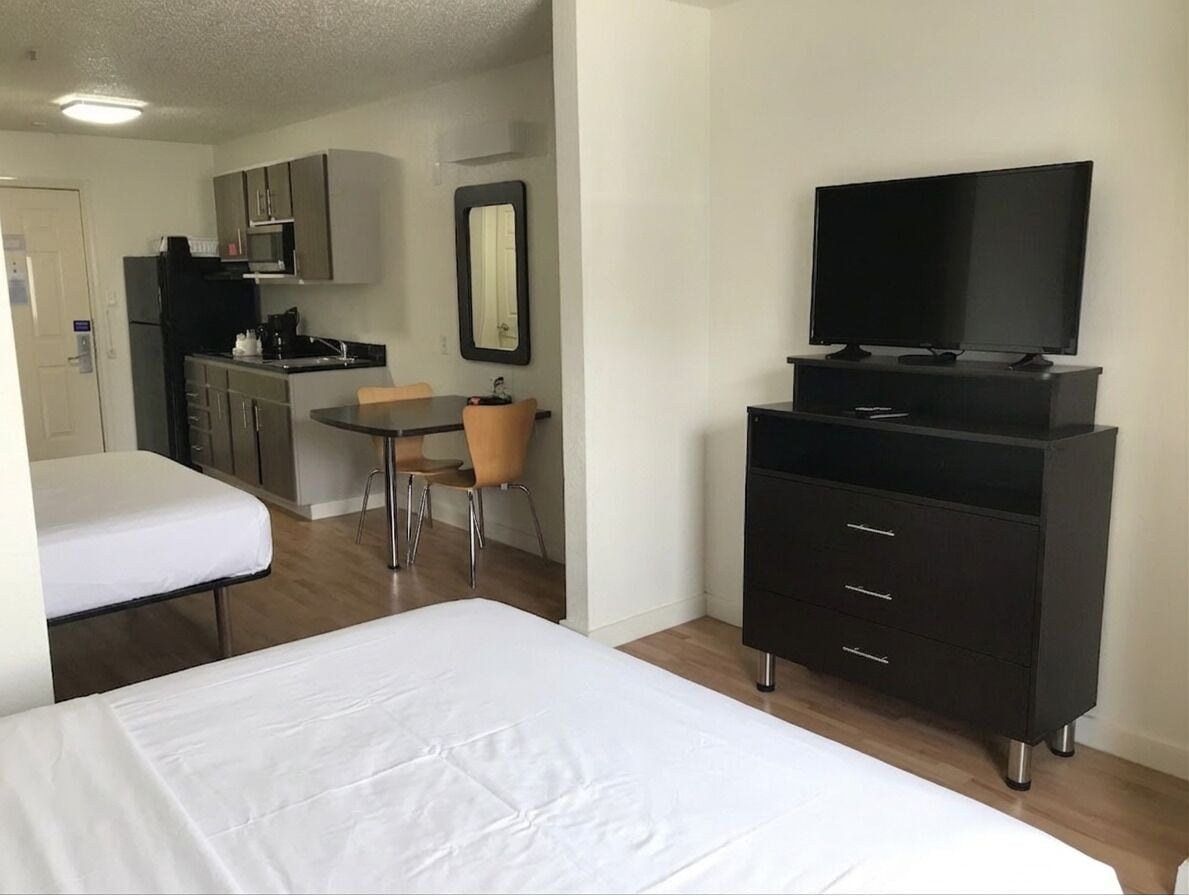 Quarto de hotel com cama, televisão, cômoda, cadeira e uma cozinha compacta.
