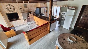 Ein gemütlicher Wohnbereich mit einer Couch, einer Holzbank, einem Esstisch mit gemustertem Tischläufer, einer Küche mit Herd, Backofen und Mikrowelle sowie einer Steinwand mit einer Uhr.