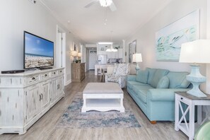 Una sala de estar con televisor, sofá, mesa de centro y una cocina al fondo.
