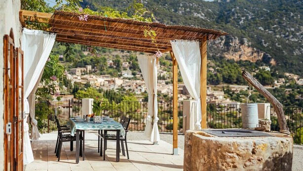 Eine Terrasse im Freien mit Tisch und Stühlen, einer Pergola mit weißen Vorhängen und Blick auf eine Stadt und Hügel im Hintergrund.