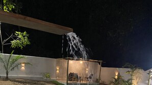 A swimming pool with a waterfall feature, surrounded by a patio with seating and wall-mounted lights. - Holiday Ranch (Japaratinga)