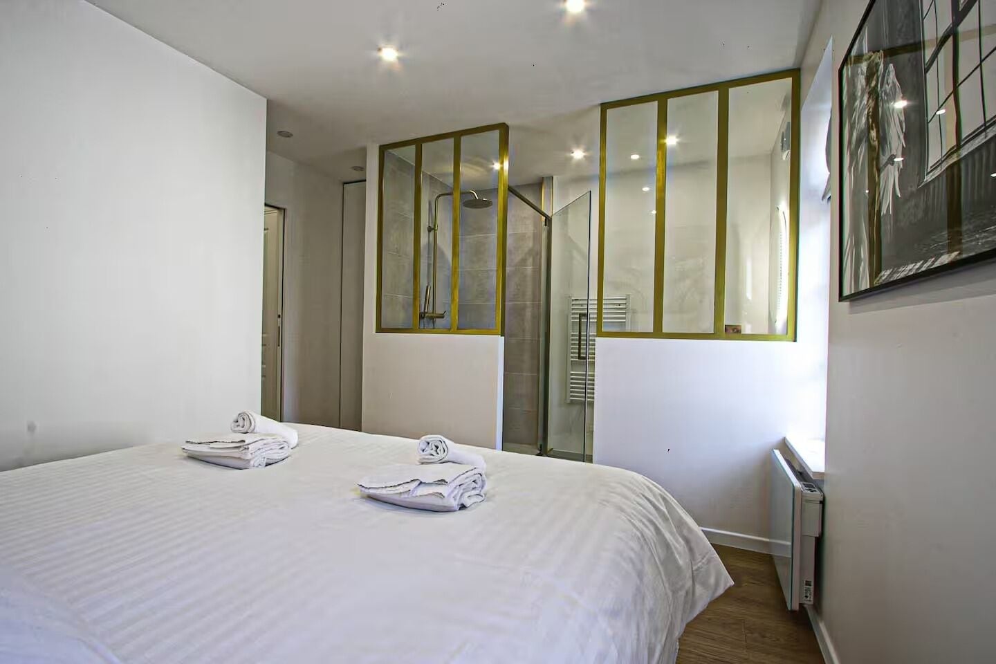 A hotel room with a large bed, a shower, a glass door, and a mirror with gold trim.