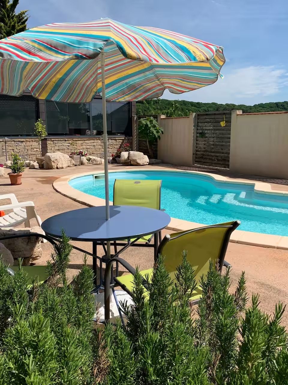 A pool area with a round table and chairs, a colorful striped umbrella, and surrounding greenery.