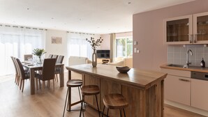 Una cocina moderna con una isla de madera, taburetes de bar, una zona de comedor con mesa y sillas, y un televisor al fondo.