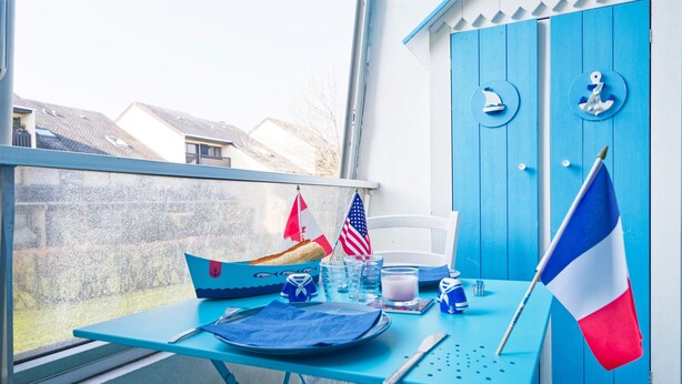 Ein Balkon mit einem Tisch für zwei Personen, mit einer französischen Flagge, einem kleinen, bootförmigen Behälter und einem Flaggenständer mit mehreren Flaggen.