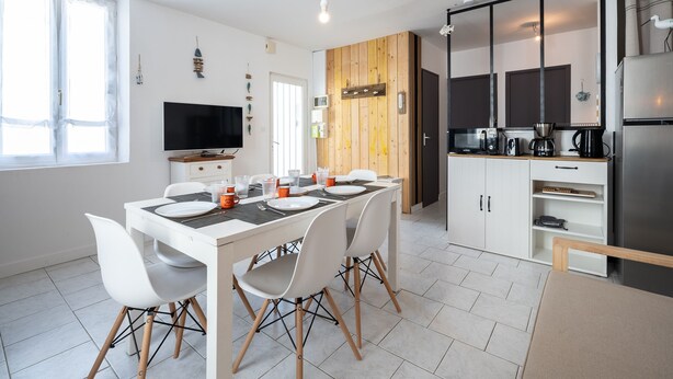 A modern kitchen with a dining area, white cabinetry, a flat-screen TV, and a wooden accent wall. - Le Refuge Loti - 200m de la Plage (Port-des-Barques)