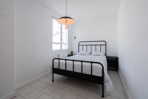Un dormitorio con estructura de cama metálica, ropa de cama blanca, una mesita de noche y una lámpara colgante, además de una ventana con cortinas traslúcidas.