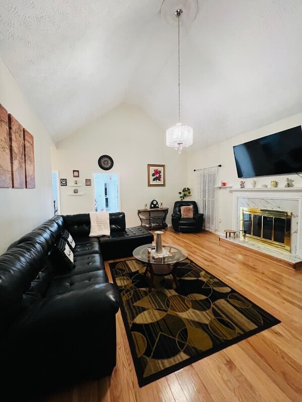 A living room with a black leather sofa, a round coffee table, a chandelier, a flat-screen TV, and a fireplace.