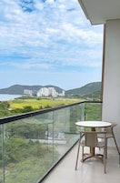 Un balcón con barandilla de vidrio, una mesa redonda y vistas a una zona costera con edificios y vegetación.